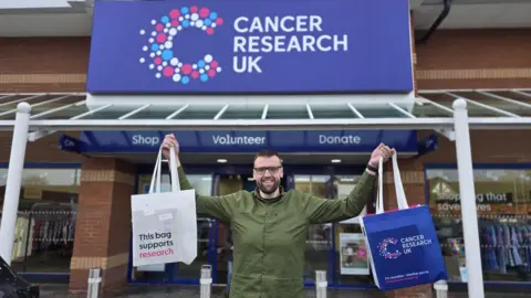 A man with dark hair, a dark beard and glasses wearing a green jacket holds up two shopping bags outside a retail space with a large blue sign that reads: "Cancer Research UK." 