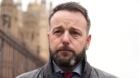 Colum Eastwood. He has short dark hair and beard, wearing a dark raincoat, white shirt and red tie.