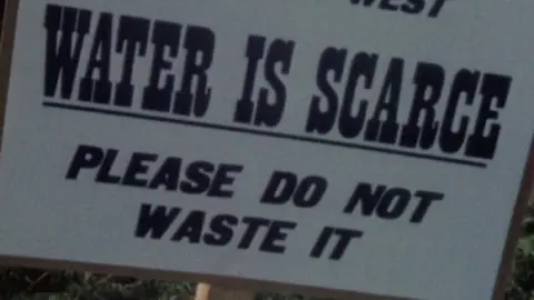 A white sign is attached to a wooden post. In bold letters it reads: "Visitors Welcome to the South West, Water is scarce. Please do not waste it."