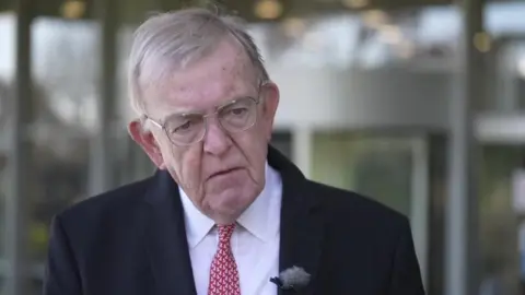 David Harris, with grey hair and glasses wearing a dark suit, white shirt, and red and white tie. 