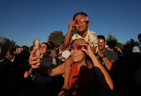A boy on an adult's shoulders holds eclipse glasses to his face in a packed crowd under a blue sky.