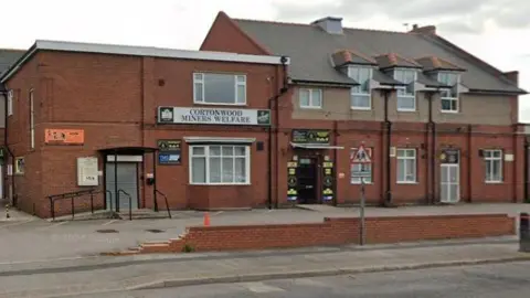A large, red-brick building viewed from across a road in daylight.
A sign above the entrance reads “Cortonwood Miners Welfare”. The main entrance has double doors and is set back slightly, with a ramp and handrails. Several additional signs and noticeboards are fixed to the brick walls near the entrances.
A triangular road sign and a low brick wall are visible along the pavement in front of the building.
The road in the foreground runs parallel to the building and has a kerb and pavement separating it from the site.