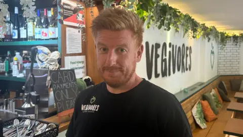 A man with ginger hair and beard wearing a black t-shirt that has white and green writing on that reads vegivores.