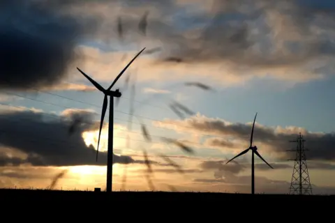 A row of still wind turbines shot through a fence. The sun is setting behind them. They are in the countryside.