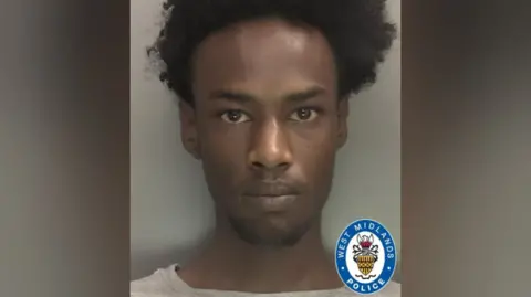 Polisi West Midlands Mugshot polisi yang menunjukkan seorang pria dengan rambut hitam keriting dan mata cokelat, mengenakan t-shirt abu-abu. Logo polisi berada di sudut kanan bawah foto.