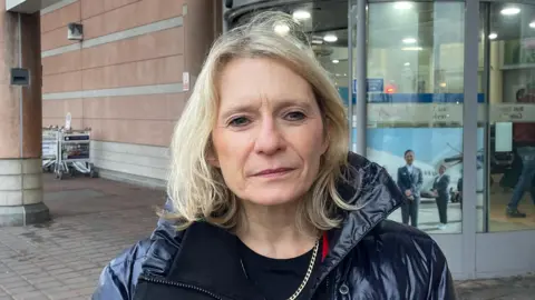 Minister Elaine Millar stood outside Jersey airport. She is wearing a navy coat above a black shirt. She is looking directly at the camera. Behind her is the entrance to the airport as well as some trolleys stacked up. 