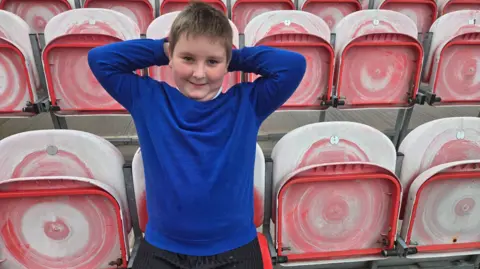 BBC Jessie sitting in the stands at the Rugby club. He has short dark hair and is wearing a navy blue jumper and has his hands behind his head. He is smiling at the camera. The seats around him are red and white.