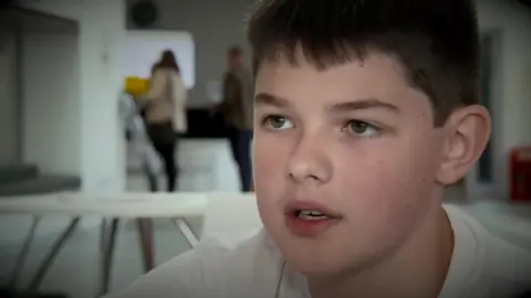 Aaron, wearing a white t-shirt, speaks to the camera in 2019