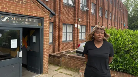 Tony Fisher A woman wearing a black dress standing outside a three storey red brick building. The entrance to her right has the address "Shire House, 400 Dallow Road" written above it.
