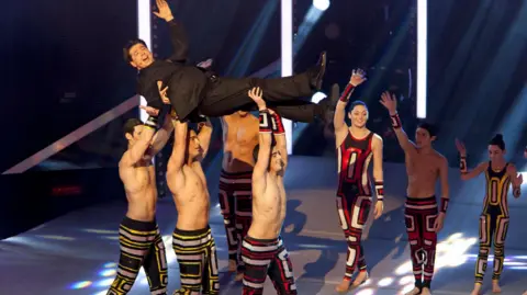 Three male acro-gymnasts from the group Spelbound carrying comedian Michael McIntyre on the Britain's Got Talent stage.