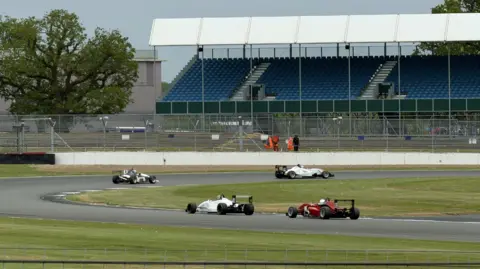 Robert Constantin/ BBC Several open-wheel race cars drive around a wide, paved racetrack during a race. In the background, there is a large grandstand with rows of blue seats and a few track staff in high-visibility clothing near the fence.