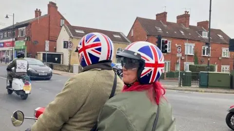 A man and a woman ride a scooter driving down the road wearing green coats and Union Jack helmets