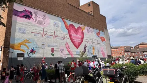 Save our Town The mural seen from further afield, with crowds of people standing below it. In the foreground are bike racks and bushes, and flats in the distance.