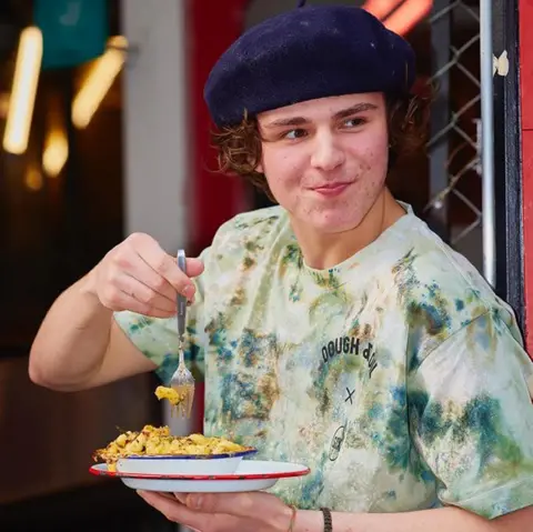 Family A young man with brown curly hair protruding from under a blue beret is wearing a T-shirt reading Dough & Oil, and holding a fork to eat from a bowl of food which he has in his other hand. 