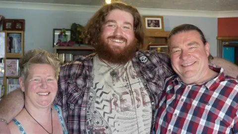 Harry Jenkins in the centre of the photo, with his mum Lorraine on his right hand side, and his dad Gary on his left-hand side. Harry has his arms around his parents shoulders and all three are smiling at the camera. They are stood in front of bookcases and photographs. Lorraine has cropped short blonde hair and is wearing a light blue floral-patterned strapped top and a black necklace. Harry has a grey patterned T-shirt and a red checked shirt on top. Harry has brown wavy hair down to his neck and a thick dark beard. Gary has pulled-back dark brown hair and is wearing a red white and blue checked shirt.