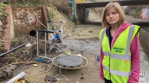 Professor Sarah Gabbott from the University of Leicester is in a hi-viz top and standing beside the Willow Brook waterway in Leicester. Alongside the channel of water, there is an array of junk and rubbish, including an old shopping trolly and a small child's trampoline. 