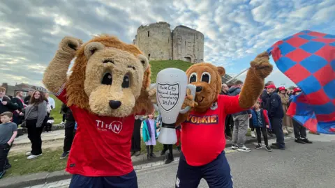 Oli Constable/BBC Two people in mascot costumes and football kits wave flags and hold an inflatable trophy