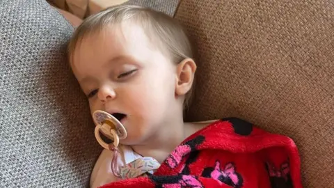 A baby girl with short blonde hair has her eyes partially closed while lying under a Red Minnie Mouse-design blanket on a sofa. She has a dummy in her mouth.