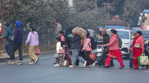 People in warm clothing, some carrying luggages and sacks, go cross a road in Kathmandu, Nepal. Cars, vans and buses are seen in the background toward the right.