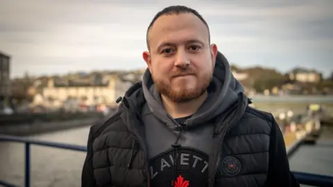 A man with short dark hair and a light coloured beard looks directly at the camera. He is standing in Bute harbour, with water, a dock and houses in the background. He is wearing a grey hooded jumper and black body warmer.