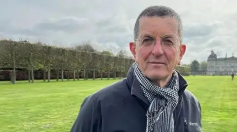 Antony Gormley is standing looking at the camera, wearing a dark grey/navy outdoor jacket and a scarf. In the distance behind him there is a large stately home.