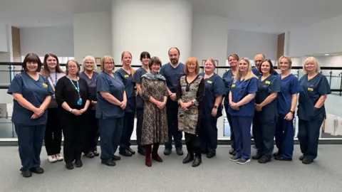 Shrewsbury and Telford Hospital Trust A large gathering of men and women in dark blue hospital uniforms standing inside a white-walled building with a grey carpet, in front of railings 