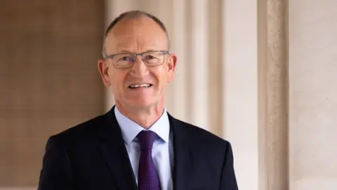 A picture of Hertfordshire County Council leader Richard Roberts wearing a black suit jacket, a purple tie, and glasses.