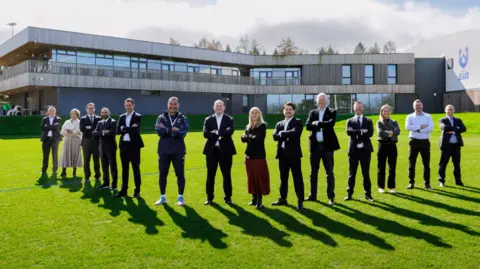 Bristol Bears A group of about a dozen people in a 'V' shape spread out on a lawn in front of a large building. They are all smiling to camera and have their arms folded. The sky is blue with puffy white clouds.