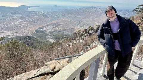 Joe Smith Seorang pria dengan celana panjang hitam, celana jins hitam dan bulu ungu berdiri di depan pagar batu dengan platform tampilan. Di belakangnya Anda dapat melihat gunung, pohon, dan laut