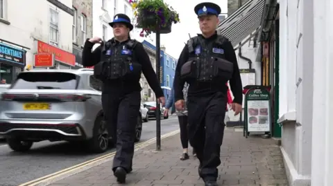 Two uniformed police officers walk side by side along a town centre pavement, passing shops and parked cars as they carry out a foot patrol.