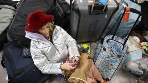 PA Media A woman, wearing a silver coat and a red hat, sits among a pile of rucksacks and suitcases as she looks at her phone