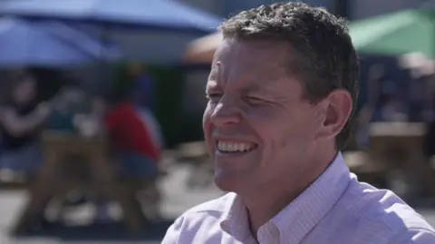Rhun ap Iorwerth squinting because the sun is bright. He is sat with a group of people on picnic benches out of focus in the background