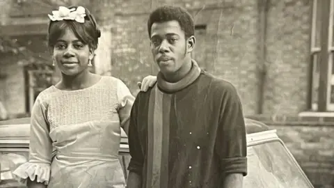 A black and white image of a man and a woman standing in front of a white car. The man is wearing a dark jumper with a stripe on the right hand side. The woman has her hand on his shoulder and is wearing a light dress and white headpiece
