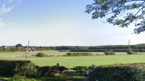 Green fields beyond a hedgerow, with a scattering of homes on the edge of the open space