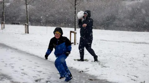 Reuters A boy dressed in a blue winter coat is smiling as he runs from a man who is about to throw a large snowball at him. There is snow on the ground and there are snowflakes in the air.