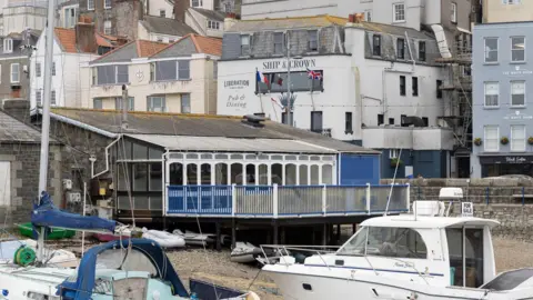 Guernsey Ports Buildings on a harbourside. There are boats moored on the beach. The building in the foreground has a balcony with a blue fence. There are houses in the background. 