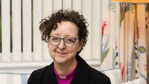 A head and shoulders image of a female bishop. She is sitting on a chair with blinds and curtains in the background. She is wearing glasses and a black blazer over a pink top with her clerical collar on. She is looking directly into the camera.