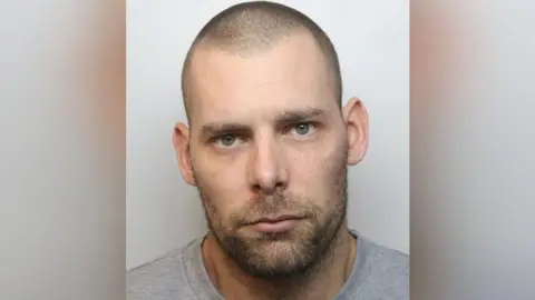 A mugshot of Damien Bendall. He has short, shaved hair and a lightly grown beard and moustache. He has green eyes and is wearing a grey T-shirt.