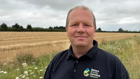 Ben Schofield/BBC Martin Lines standing on the edge of a freshly harvested wheat field. He is wearing a dark blue collared T-shirt with the logo of the Nature Friendly Farming Network on his chest. He is partly bald and has short light brown hair. Behind him is a strip of wild flowers and grasses, which borders a wheat field. In the further distance is a mature hedgerow.