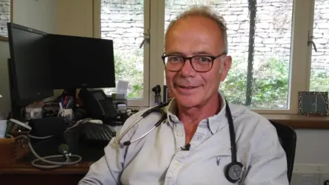 Dr Simon Opher sat in a room at a doctors surgery with a stethoscope around his neck