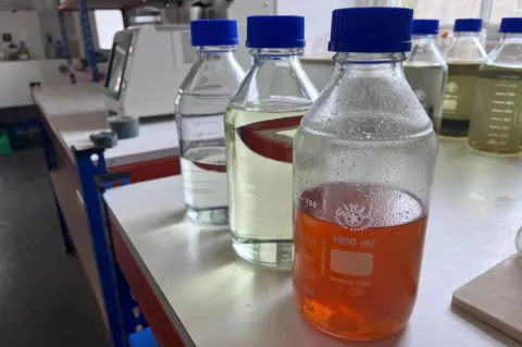 Three bottles on a lab worktop, each with a blue cap. The closest is a darker liquid with solids floating in it. The other two are transparent liquids.