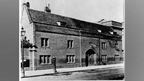 Museum of Cambridge A black and white photograph of a now-demolished building known as the Spinning House in Cambridge. It is brick built with mullioned windows on its ground and first storeys. There are small dormer buildings in its pitched roof. It has a large arched doorway with a door made of wooden planks.
