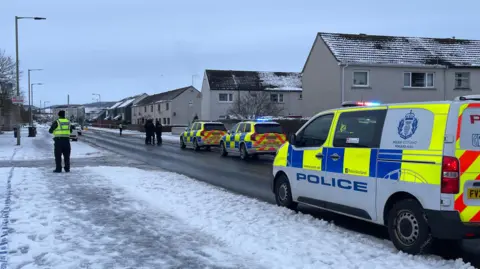 A police van and an officer in a hi-vis vest over black clothing standing next to a snow path in Inverness.