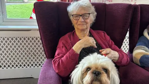 Phil Gorman An elderly woman with grey hair and black-rimmed glasses is pictured sitting in a purple high-backed armchair. She is wearing a burgundy pink cardigan and there is a fluffy-haired cream-coloured dog in front of her. In the background there is a white radiator cover below a window.