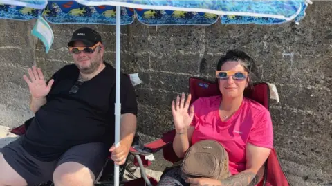 A picture of two people sat on a beach waving at the camera.