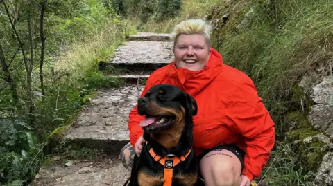 Rowena Halstead Rowena Halstead, a woman wearing a red coat, is crouching alongside a dog. She has short blonde hair and is smiling for the photo. There are stone steps behind her.