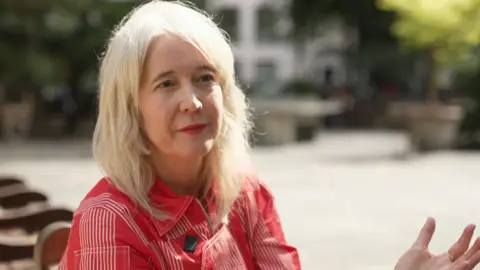 A woman with blonde hair, red lipstick and a red a red and white striped dress sits on a park bench in the sunshine