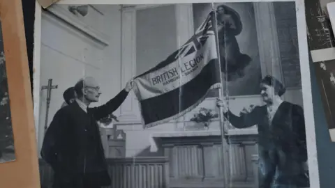 Eddie Wulff In another black and white photograph, Mabel Wulff is pictured on the right holding the British Legion flag aloft in the English Church in Hamburg. Pictured with two men in suits, the trio are holding the flag up in front of the altar. 