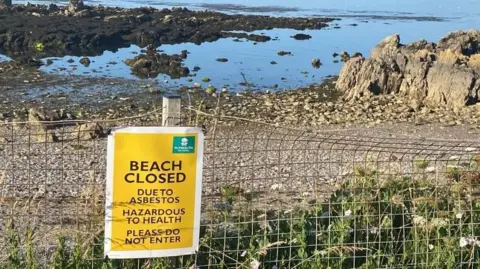 An empty Mount Batten beach near Plymouth which has been closed following the discovery of asbestos. There is a yellow warning signing saying "BEACH CLOSED". The beach is stoney with lots of brown seaweed on it. The water is dark blue coloured with bright blue skies above it. 