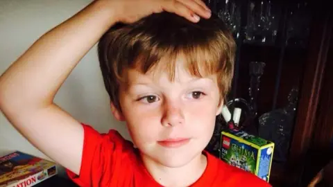Anna-Louise Bates A young boy with short brown hair is smiling. He has one hand on his head. Behind him is a box of Lego and a board game.